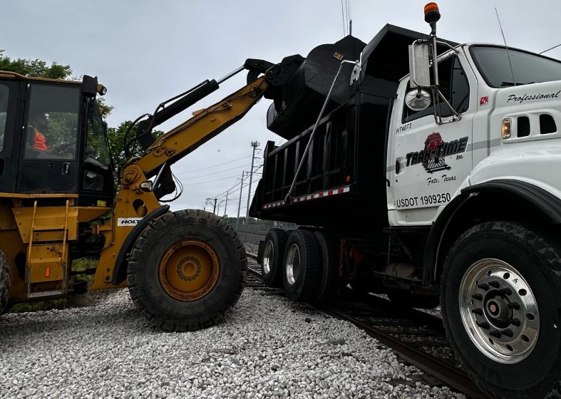 A yellow loader lifts debris into a white dump truck on gravel.