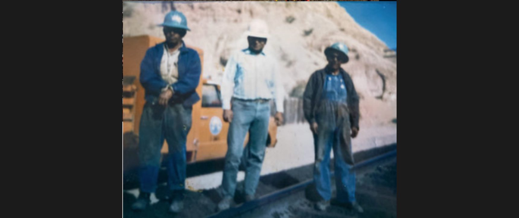 Three men standing by a road with a rocky hill in the background.